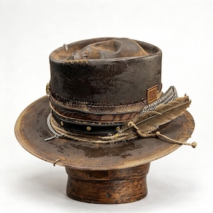 Könnte beinhalten: Ein dunkelbrauner, abgenutzter Fedora-Hut mit strukturierter Oberfläche, einem schwarzen Lederband mit goldfarbenen Nieten, einer silbernen Kette und einer natürlichen Feder in einer geflochtenen Borte. Ein kleines quadratisches Lederetikett ist seitlich angebracht.