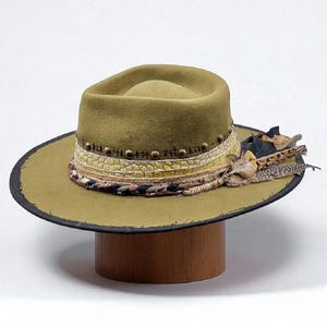 Puede incluir: Sombrero fedora verde oliva con ala ancha y banda decorativa. La banda presenta un patrón texturizado, detalles de tachuelas y un adorno de plumas. El sombrero se exhibe sobre un soporte de madera sobre un fondo blanco.