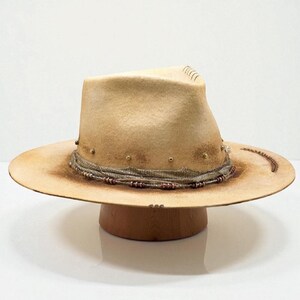 May include: A tan-coloured fedora hat with a wide brim, decorated with a band and small gold-coloured studs. The hat is displayed on a wooden hat stand, highlighting its shape and details. The brim has a distressed appearance.