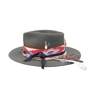 Könnte beinhalten: Dunkelgrüner Fedora-Hut mit breiter Krempe. Der Hut ist mit einem rot-weiß-blauen gemusterten Band, einem marineblauen Band, Perlen und Federn verziert. Ein kleines Muschel- und Sternchen-Charm ist an dem Hut befestigt.