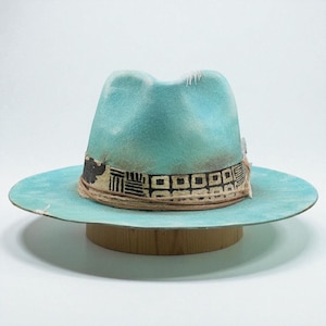 May include: A turquoise fedora hat with a wide brim. The hat has a decorative band with a geometric pattern and a feather accent. The hat is displayed on a wooden hat stand against a white background.