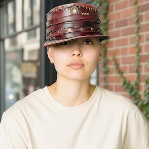 Peut inclure: Un chapeau en cuir bordeaux avec un bord plat et une couronne multicouche. Le chapeau présente des coutures décoratives, des œillets métalliques et un petit ornement sur le dessus. Le chapeau est porté par une personne portant un t-shirt crème.