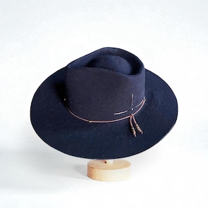 May include: A navy blue fedora hat with a wide brim, displayed on a wooden hat stand. The hat features a brown leather band with decorative beads and a tassel. The hat's crown has a slight indentation.