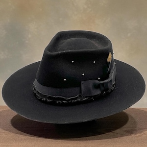 May include: A black fedora hat with a wide brim. The hat has a black ribbon with a bow, decorative trim, and small silver studs. A feather is tucked into the ribbon. The hat is displayed on a wooden surface.