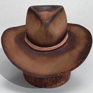 May include: A brown cowboy hat with a wide brim and a pinched crown. The hat has a light brown band around the base of the crown. The hat is made of a material that looks like leather or a leather-like material. The hat is on a wooden base.