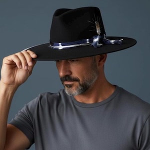 Può includere: Un cappello fedora nero con tesa larga. Il cappello presenta un nastro blu e bianco ed è adornato da piume. Il cappello è tenuto da una persona che indossa una maglietta grigia.
