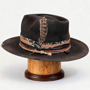 Puede incluir: Un sombrero fedora marrón oscuro con ala ancha, adornado con una banda estampada, detalles de cuero y una pluma decorativa. El sombrero se exhibe en un soporte de madera.