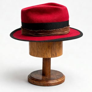 Może przedstawiać: Czerwony kapelusz typu fedora z czarną wstążką i brązowym wykończeniem, prezentowany na drewnianym stojaku na kapelusze. Kapelusz ma klasyczny kształt z lekko wklęsłą koroną i szerokim rondem. Stojak jest drewniany.
