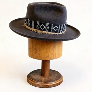 Könnte beinhalten: Ein anthrazitfarbener Fedora-Hut mit einem dekorativen Band mit schwarz-weißem geometrischen Muster. Der Hut wird auf einem hölzernen Hutständer präsentiert. Der Hut hat eine Krempe von etwa 7.6 cm Breite.