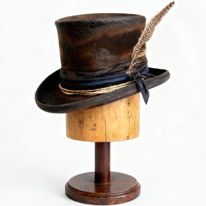 Può includere: Un cappello a cilindro marrone con una piuma, appoggiato su un espositore in legno. Il cappello presenta un nastro blu scuro e un bordo simile a una corda. L'espositore è in legno e ha una base rotonda.