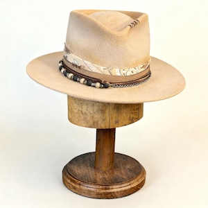 May include: A beige fedora hat with a wide brim, decorated with a patterned band, beads, and a chain. The hat is displayed on a wooden hat stand. The hat's brim is approximately 35.5 cm in diameter.
