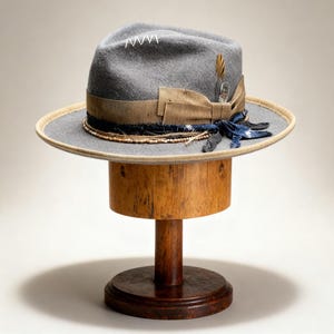 Könnte beinhalten: Ein grauer Fedora-Hut mit breiter Krempe, verziert mit einem beigen Band, einer Feder und einem Denim-Akzent. Der Hut wird auf einem hölzernen Hutständer mit rundem Sockel und zylindrischem Oberteil präsentiert.