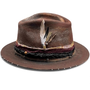 Könnte beinhalten: Ein brauner Fedora-Hut mit einem Used-Look. Der Hut hat ein dekoratives Band mit schwarzen, bordeauxroten und goldenen Akzenten und ist mit Federn verziert. Der Rand ist mit kleinen silbernen Nieten versehen, was eine rustikale, Vintage-Ästhetik erzeugt.
