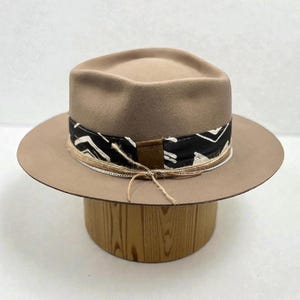 Op de afbeelding: Een bruin fedora hoed met een zwart-wit patroon band en een bruin lederen accent. De hoed heeft een brede rand en wordt getoond op een houten hoedenstandaard. De band is geaccentueerd met een dun touw en vastgebonden met touw.