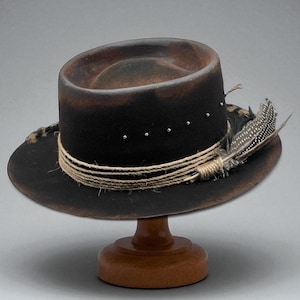 Puede incluir: Un sombrero estilo fedora marrón oscuro con un acabado envejecido, adornado con una banda de cuerda y plumas. El sombrero presenta pequeños tachuelas plateadas y se apoya en un soporte de madera. El ala del sombrero tiene unos 35.5 cm de diámetro.