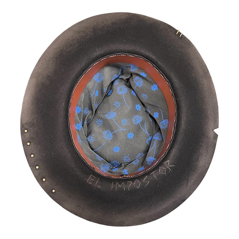 May include: A dark brown hat with a wide brim, viewed from the inside. The interior features a blue floral patterned lining, a brown leather band, and the text "EL IMPOSTOR". Gold-coloured studs are visible around the brim.