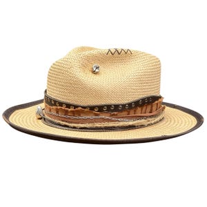 Puede incluir: Sombrero fedora de paja con ala marrón y banda decorativa. La banda presenta cuero marrón, tachuelas metálicas y una cadena. Un dije plateado y una pequeña campana están adheridos al sombrero.