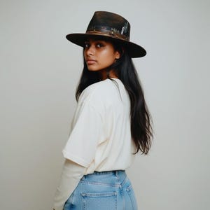 May include: A dark brown fedora hat with a wide brim. The hat features a leather band with decorative metal accents and feathers. The hat is worn by a person wearing a white long-sleeve shirt and blue jeans.