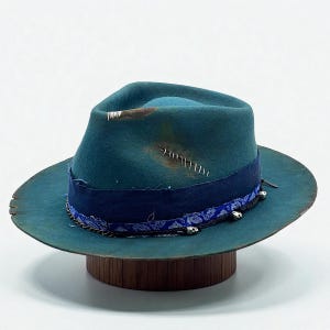 Könnte beinhalten: Türkisfarbener Fedora-Hut mit Used-Look. Der Hut hat ein dunkelblaues Band mit silbernen Akzenten und einen Paisley-Besatz. Der Hut wird auf einem Holzständer vor weißem Hintergrund präsentiert.