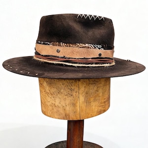 May include: A dark brown fedora hat with a wide brim. The hat has a decorative band with leather and patterned fabric accents, and is decorated with small metal studs. The crown has white stitching.