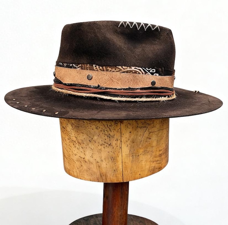 May include: A dark brown fedora hat with a wide brim. The hat has a decorative band with leather and patterned fabric accents, and is decorated with small metal studs. The crown has white stitching.