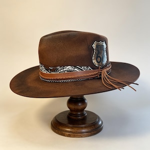 Puede incluir: Un sombrero fedora de fieltro marrón con un emblema plateado de la Ruta 66 en el lateral. El sombrero está adornado con un pañuelo de patrón blanco y negro, un cordón de cuero trenzado, una cadena metálica y varios flecos de cuero colgantes.