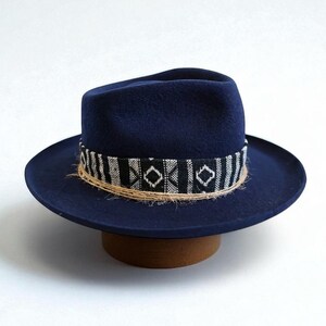Pode incluir: Um chapéu fedora azul marinho com uma faixa decorativa com um padrão geométrico em preto e branco. O chapéu tem uma aba larga e é exibido em um suporte de chapéu marrom. A faixa é feita de material tecido.