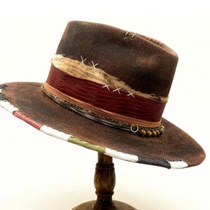 Könnte beinhalten: Ein brauner Fedora-Hut mit breiter Krempe, verziert mit einem roten Band und dekorativen Nähten. Der Rand ist mit einem Muster aus roten, weißen, grünen und schwarzen Streifen versehen. Der Hut wird auf einem Holzständer präsentiert.