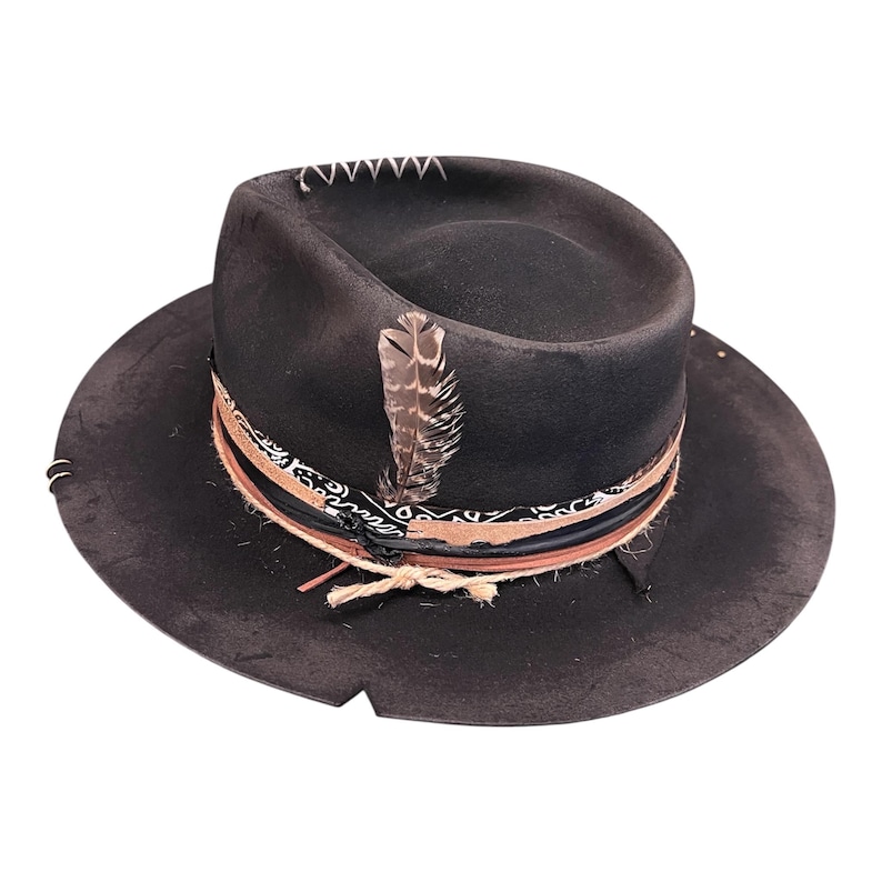 May include: A black fedora hat with a wide brim, decorated with a feather, patterned band, and rope detailing. The hat has white stitching on the crown and a distressed finish, giving it a vintage look.
