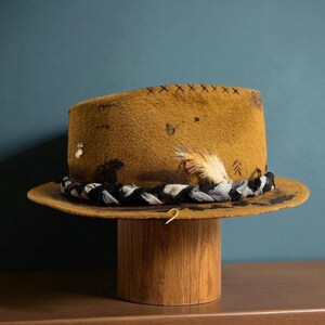 May include: Mustard-coloured fedora hat with a distressed finish, featuring a decorative braided band in black and white. The hat is adorned with a feather and small embellishments. It is displayed on a wooden hat stand.