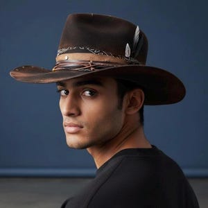 May include: A brown cowboy hat with a wide brim and a decorative leather band. The hat features a feather accent and a distressed finish, giving it a vintage look. The hat is a stylish accessory.