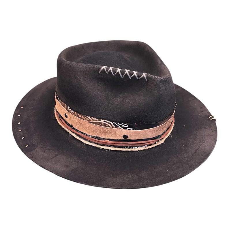 May include: A black fedora hat with a wide brim and distressed finish. The hat has a decorative band with patterned fabric, leather, and rope detailing. Silver studs are placed around the brim. White stitching is visible on the crown.