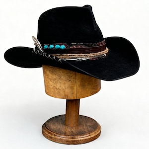 May include: A black cowboy hat with a wide brim, adorned with a patterned band, turquoise beads, a feather, and a leather cord. The hat is displayed on a wooden hat stand, showcasing its shape and details. The hat's design suggests a Western or bohemian style.