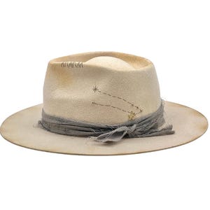 Op de afbeelding: Crèmekleurige fedora hoed met een brede rand. De hoed heeft een grijze stoffen band met een geknoopt detail en een sterrenbeeldontwerp met goudkleurige steraccenten. De hoed heeft een distressed, vintage look.