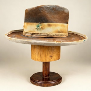 Puede incluir: Un sombrero fedora marrón con ala ancha y una banda de color claro. El sombrero se exhibe en un soporte de sombrero de madera. La banda del sombrero presenta un diseño cosido en verde. El ala tiene un ribete de color claro.