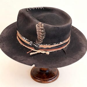 May include: A dark grey fedora hat with a wide brim, decorated with a patterned band, leather details, and a feather. The hat has white stitching on the crown and is displayed on a wooden hat stand.