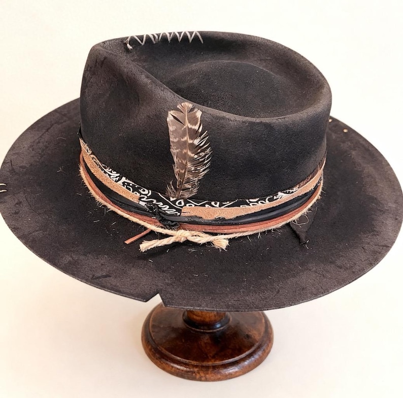 May include: A dark grey fedora hat with a wide brim, decorated with a patterned band, leather details, and a feather. The hat has white stitching on the crown and is displayed on a wooden hat stand.