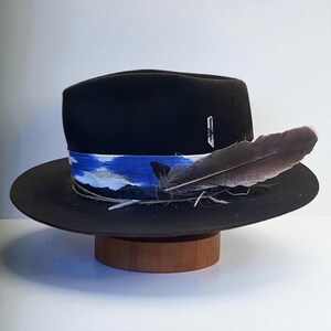 Pode incluir: Um chapéu fedora preto com aba larga, com uma faixa estampada azul e branca e uma pena grande. O chapéu é exibido em um suporte de madeira, mostrando seu design e artesanato. Um alfinete prateado está na lateral do chapéu.