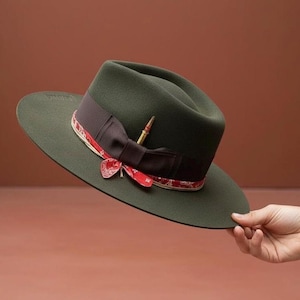 Könnte beinhalten: Ein dunkelgrüner Fedora-Hut mit breiter Krempe. Der Hut hat ein dunkelbraunes Band mit rot-weißem Muster. Eine goldfarbene Nadel ist befestigt. Der Hut wird von einer Hand gehalten.