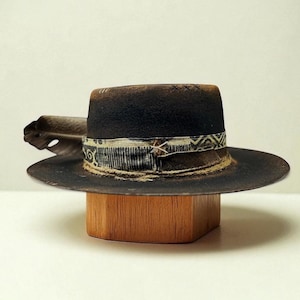 May include: A distressed black fedora hat with a wide brim, featuring a patterned band and a feather accent. The hat is displayed on a wooden block, showcasing its vintage style and craftsmanship. The hat's brim measures approximately 7.6 cm.