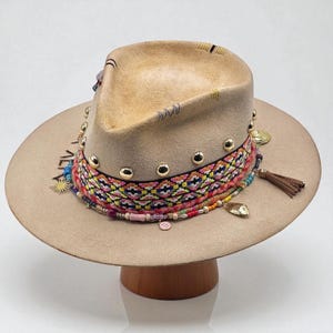 Può includere: Un cappello fedora color beige con tesa larga. Il cappello presenta una fascia intrecciata colorata, borchie dorate e ciondoli decorativi. Una piccola nappa pende dal lato. Il cappello è esposto su un supporto di legno.