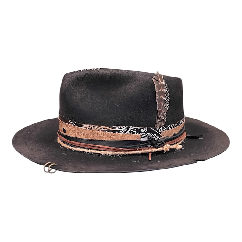 May include: Black fedora hat with a wide brim. The hat features a patterned band, a brown leather band, and a rope band. A decorative feather is attached to the side. Two silver rings are attached to the brim.