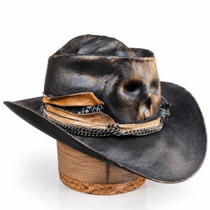 May include: A black cowboy hat with a distressed finish and a moulded skull detail on the front crown. The hat features a layered band consisting of a tan leather strip, a black and white patterned fabric, and a silver-toned metal chain.