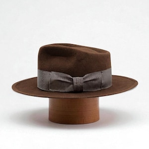 May include: A brown fedora hat with a grey ribbon band and bow, displayed on a wooden hat stand. The hat has a wide brim and a classic shape. The hat stand is cylindrical.