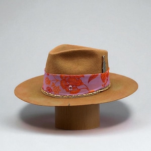 May include: A brown fedora hat with a wide brim, adorned with a pink and orange floral patterned band. The band is trimmed with gold beading. A small feather and jewel accent the hat.