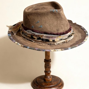 Puede incluir: Un sombrero fedora marrón con ala ancha, adornado con bandas de tela decorativas en varios patrones y colores. El sombrero presenta costuras azules y se exhibe en un soporte de sombrero de madera.
