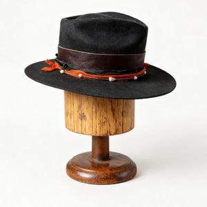 May include: A black fedora hat with a brown leather band and orange ribbon, decorated with small white beads. The hat is displayed on a wooden hat stand with a round base and cylindrical top, against a white backdrop.