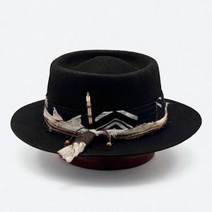 Peut inclure: Un chapeau fedora noir avec un large bord. Le chapeau est orné d'une bande à motifs avec des motifs géométriques blancs et noirs, ainsi que d'un accent de plumes et de perles. Le chapeau est présenté sur un support en bois foncé.