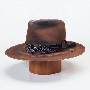 Op de afbeelding: Een verweerde bruine fedora hoed met een brede rand en een zwart-witte paisley band. De hoed heeft een donkerbruine kroon en een decoratief zwart lint. De hoed wordt op een houten standaard gepresenteerd.