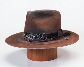 Może przedstawiać: Zniszczony brązowy kapelusz typu fedora z szerokim rondem i czarno-białą opaską w paisley. Kapelusz ma ciemnobrązową koronę i ozdobną czarną wstążkę. Kapelusz jest prezentowany na drewnianym stojaku.